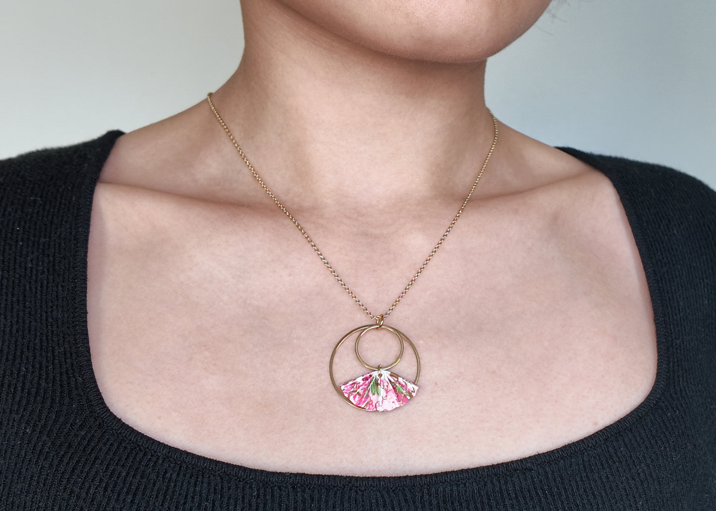 A gold-colored necklace with a double hoop fan-shaped pendant made of pink origami paper displayed on a model